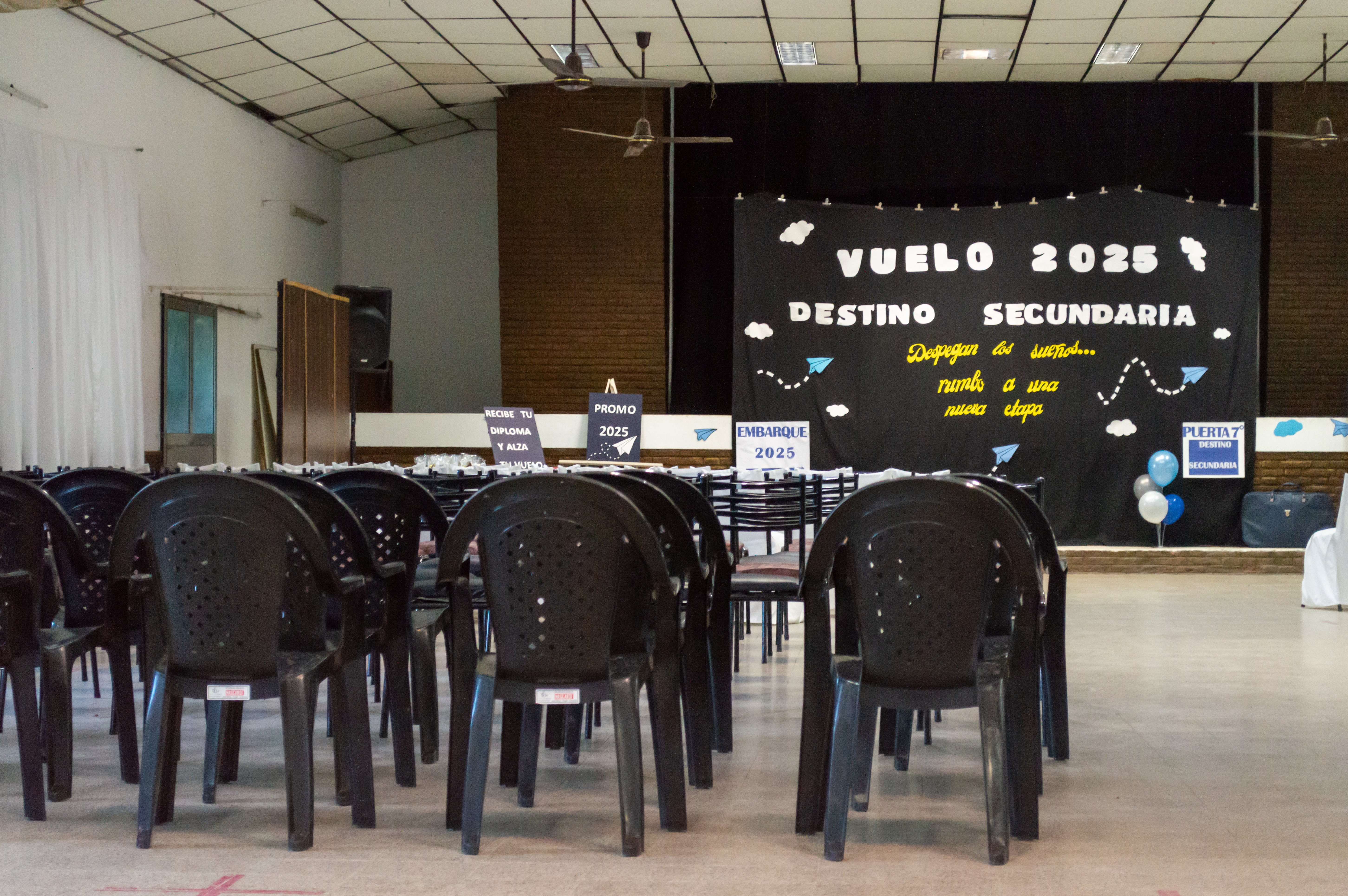 Salón preparado para un acto escolar, con filas de sillas frente a un espacio decorado con el cartel. Soldini.