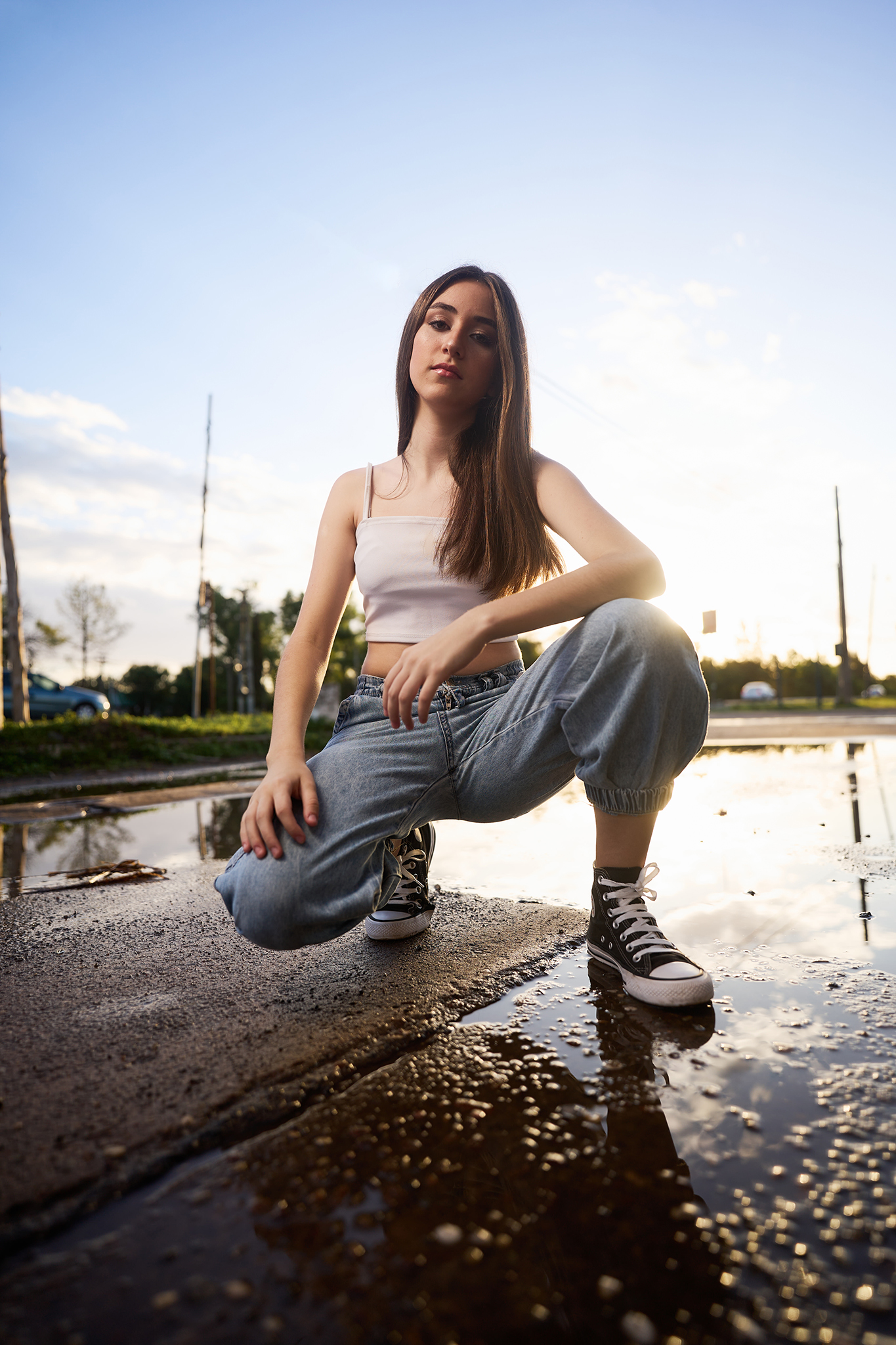 Pose en cuclillas sobre un charco que refleja el cielo, a contraluz del atardecer. Book de quince al aire libre de DNX Estudio, Funes, Rosario.