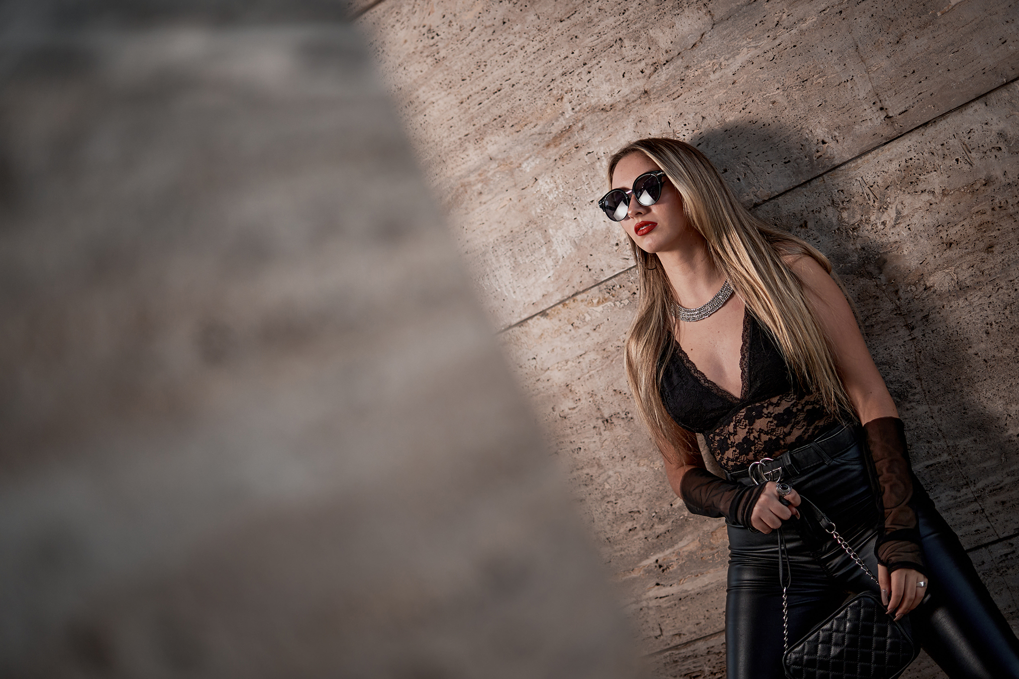 Retrato de moda contra una pared de travertino: lentes de sol, encaje negro y pantalón de cuero. Producción urbana de DNX Estudio, Funes, Santa Fe.