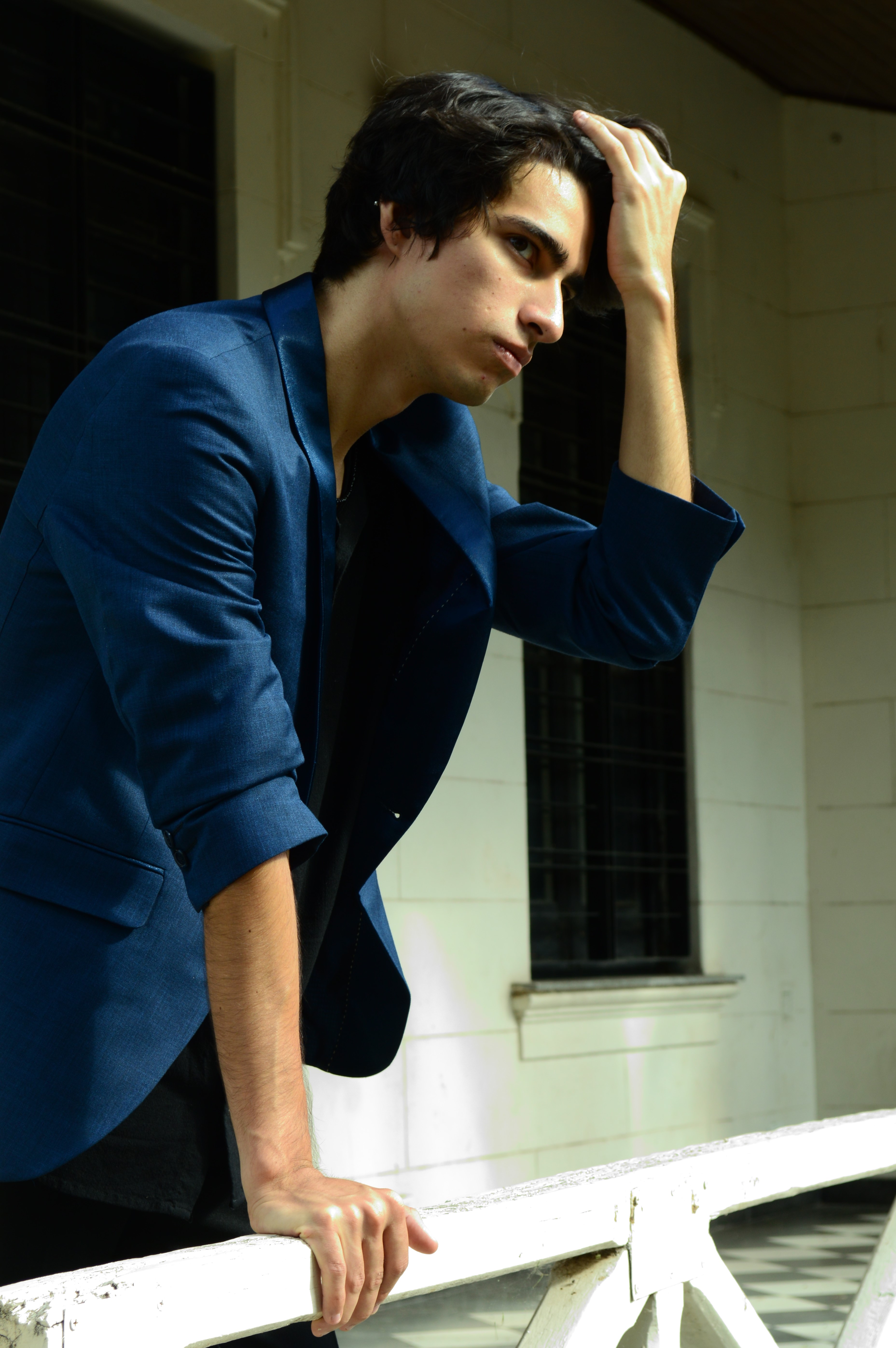 Un hombre con traje azul oscuro posa apoyado en una baranda junto a una ventana, en un retrato editorial de moda con luz natural. Rosario.