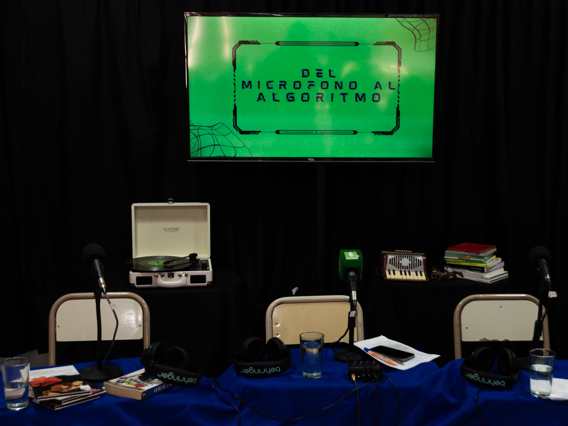 Espacio de exposición con mesas, objetos y equipos, frente a una pantalla verde con letras negras durante una jornada institucional. Rosario.