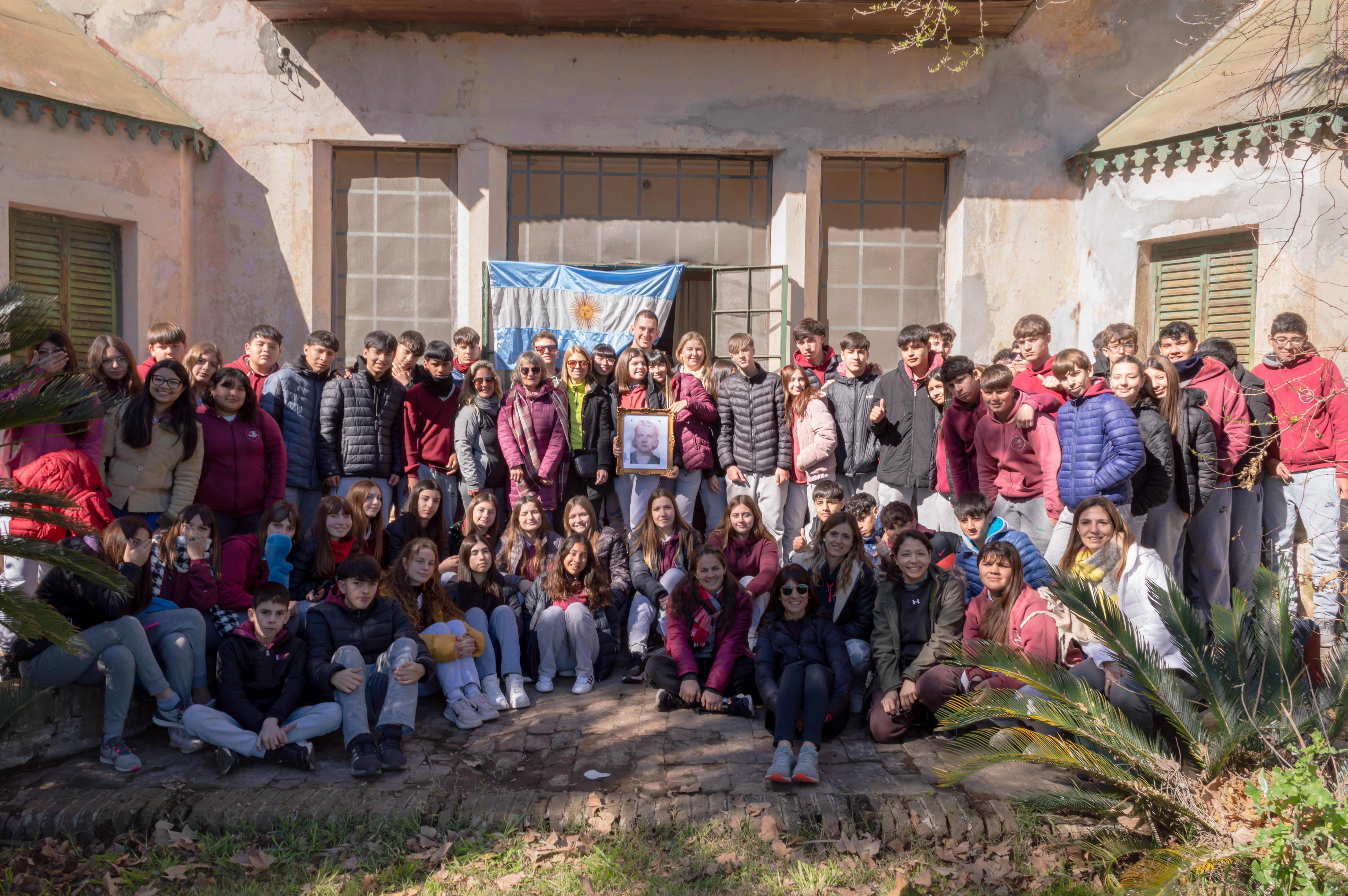 Un grupo numeroso posa frente a un edificio de ladrillo durante una salida, rodeado de árboles y vegetación. Soldini.