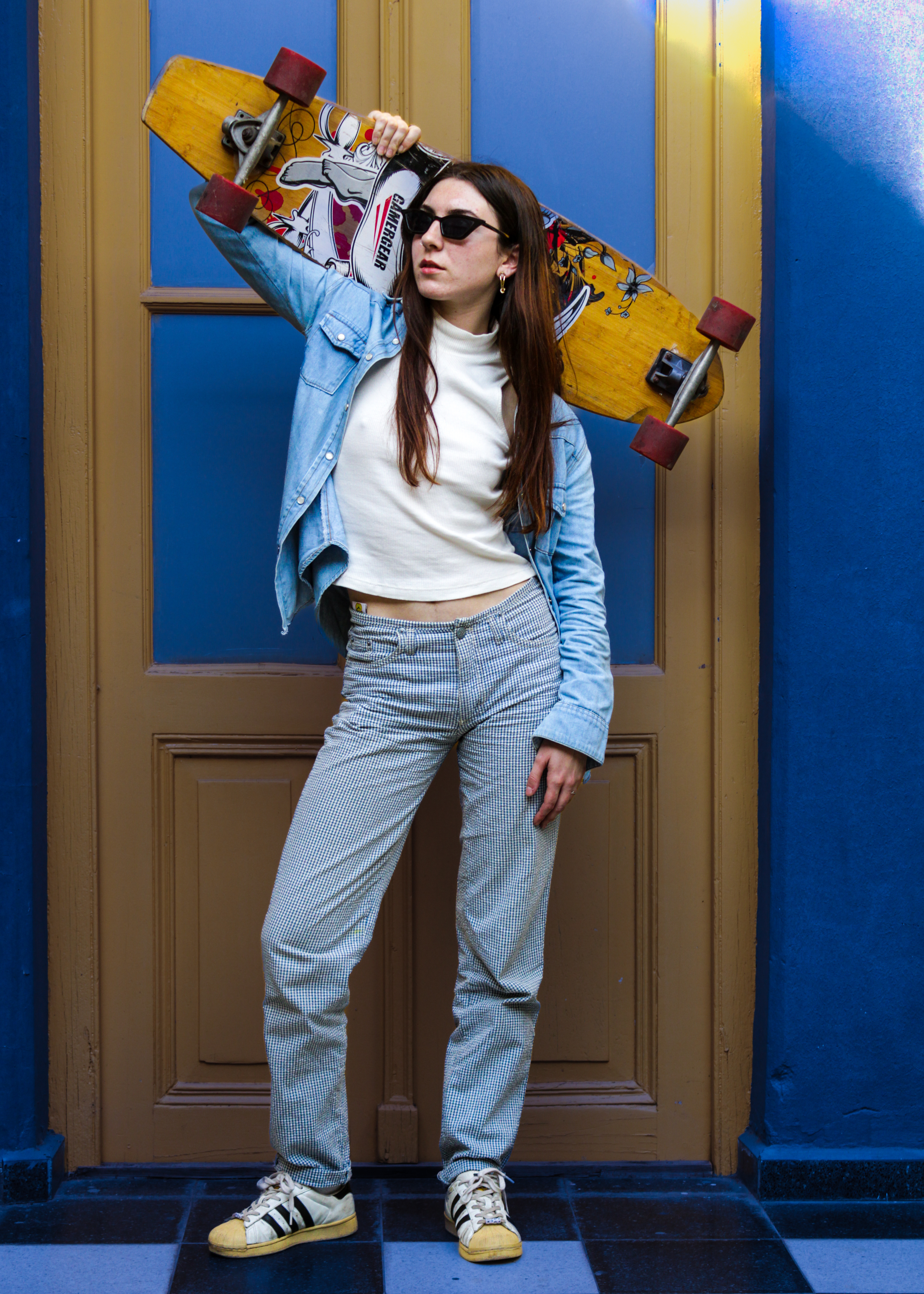 Una mujer con gafas de sol, remera clara y pantalón celeste posa frente a una puerta azul y amarilla en un entorno urbano. Rosario.