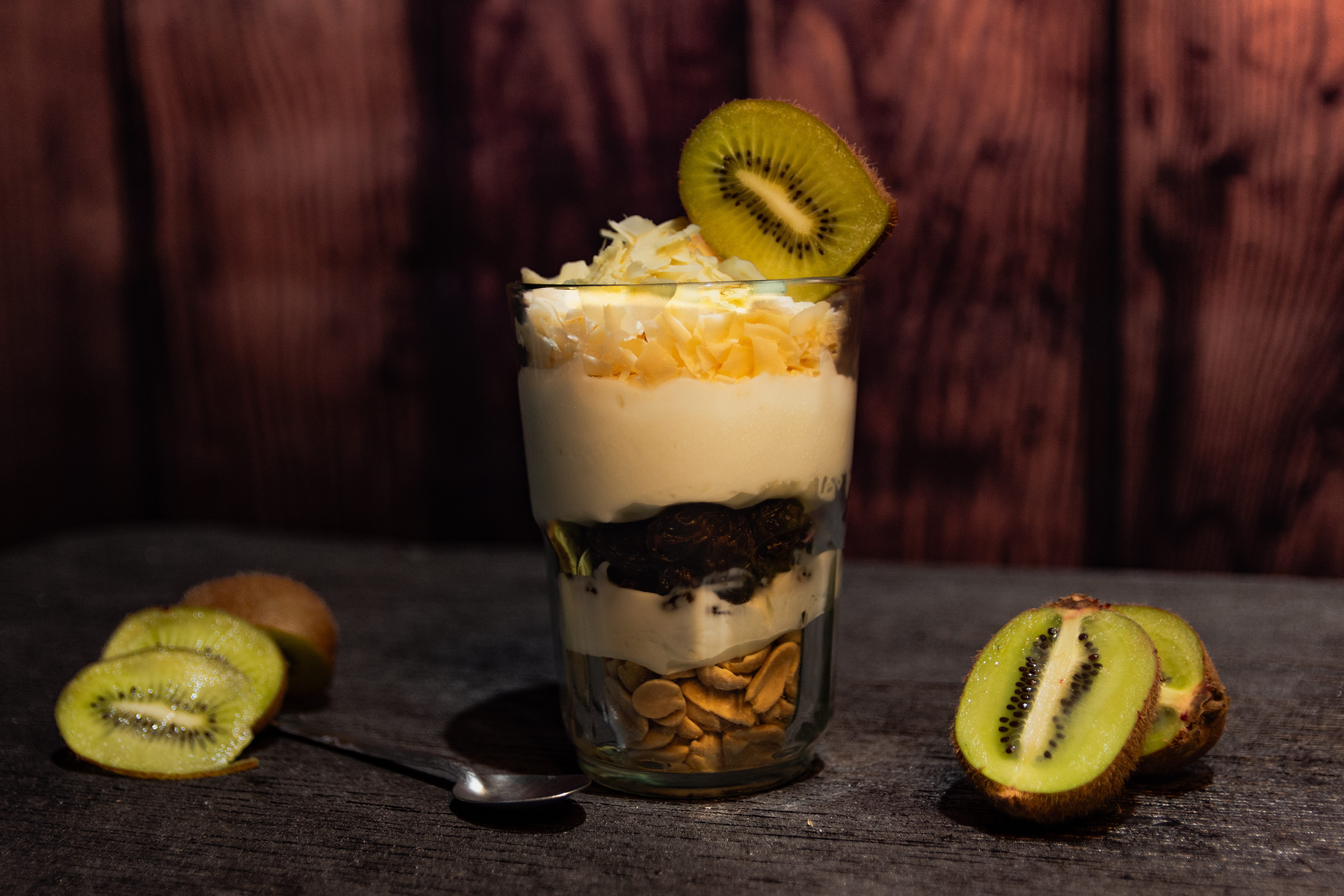 Postre en vaso transparente, con capas de yogur y kiwi, coronado con crema. Acompañado de rodajas y mitades de kiwi sobre una mesa oscura. San Lorenzo.
