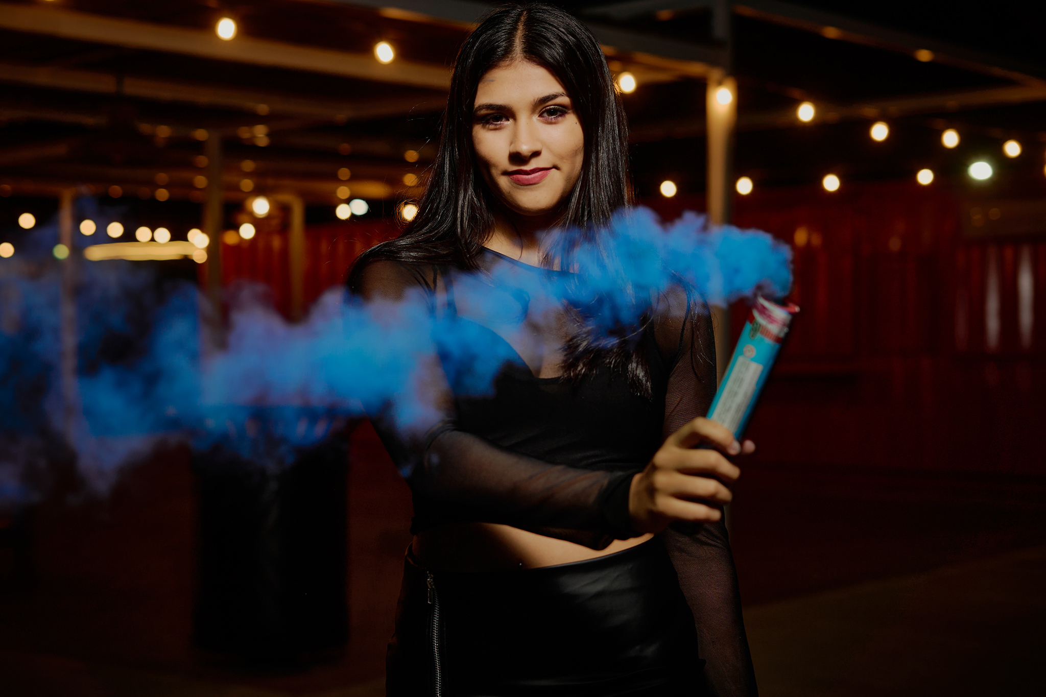 Chica de negro sosteniendo una bengala de humo azul, bajo guirnaldas de luces. Sesión de 15 con humo de color de DNX Estudio, Funes, Rosario.